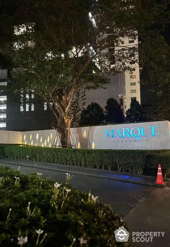 Elegant night view of Marque Sukhumvit entrance with illuminated signage and lush greenery.