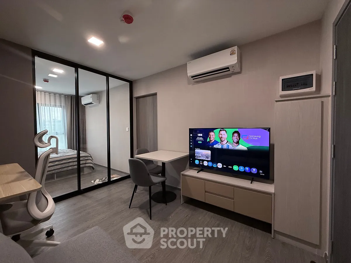 Modern living room with sleek furniture and large TV, adjacent to a cozy bedroom with glass partition.