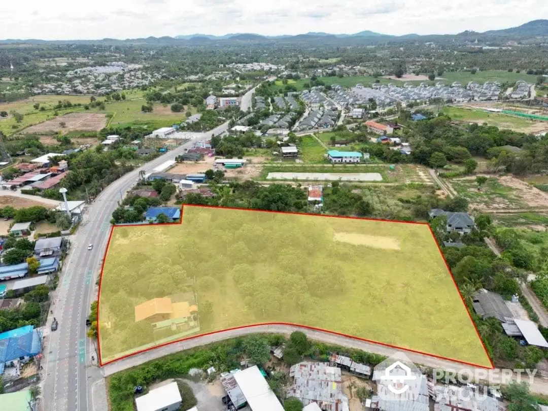 Expansive aerial view of a large, undeveloped land parcel outlined in red, nestled in a vibrant community with road access and scenic backdrop, ideal for development.