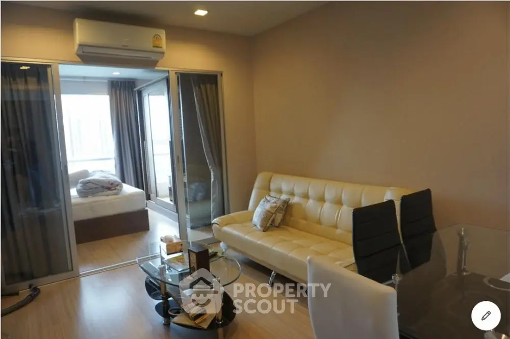 Modern living room with glass table, beige sofa, and view into bedroom, featuring sleek design and cozy ambiance.