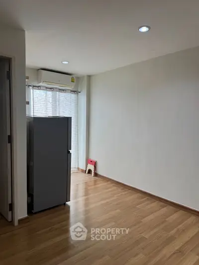 Spacious unfurnished room with wooden flooring and natural light, featuring a refrigerator and air conditioning.