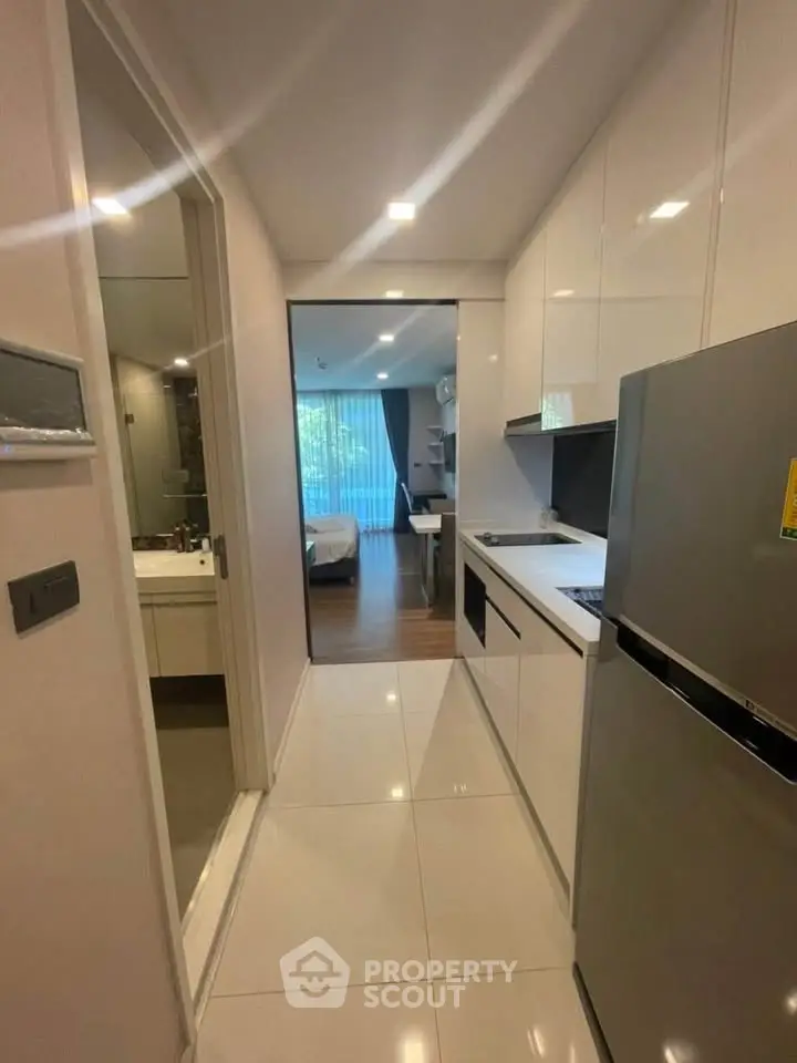 Modern apartment kitchen with sleek white cabinets and stainless steel fridge, leading to a cozy bedroom.