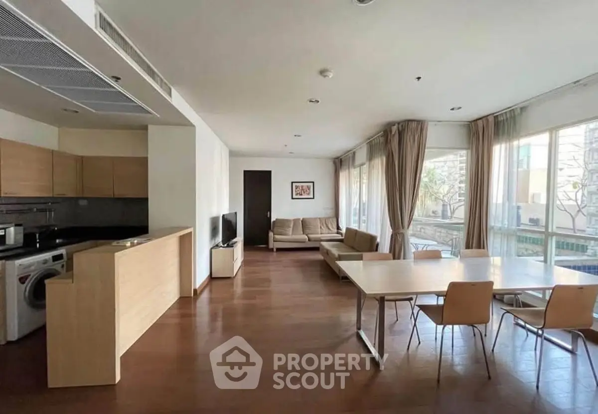 Spacious modern living room with open kitchen and dining area, featuring large windows and wooden flooring.