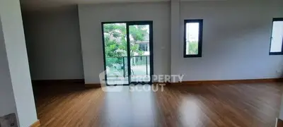 Spacious empty room with wooden flooring and large windows overlooking greenery.