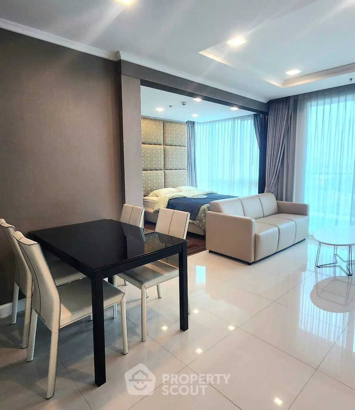 Modern open-plan living room with dining area and bedroom, featuring sleek furniture and large windows.