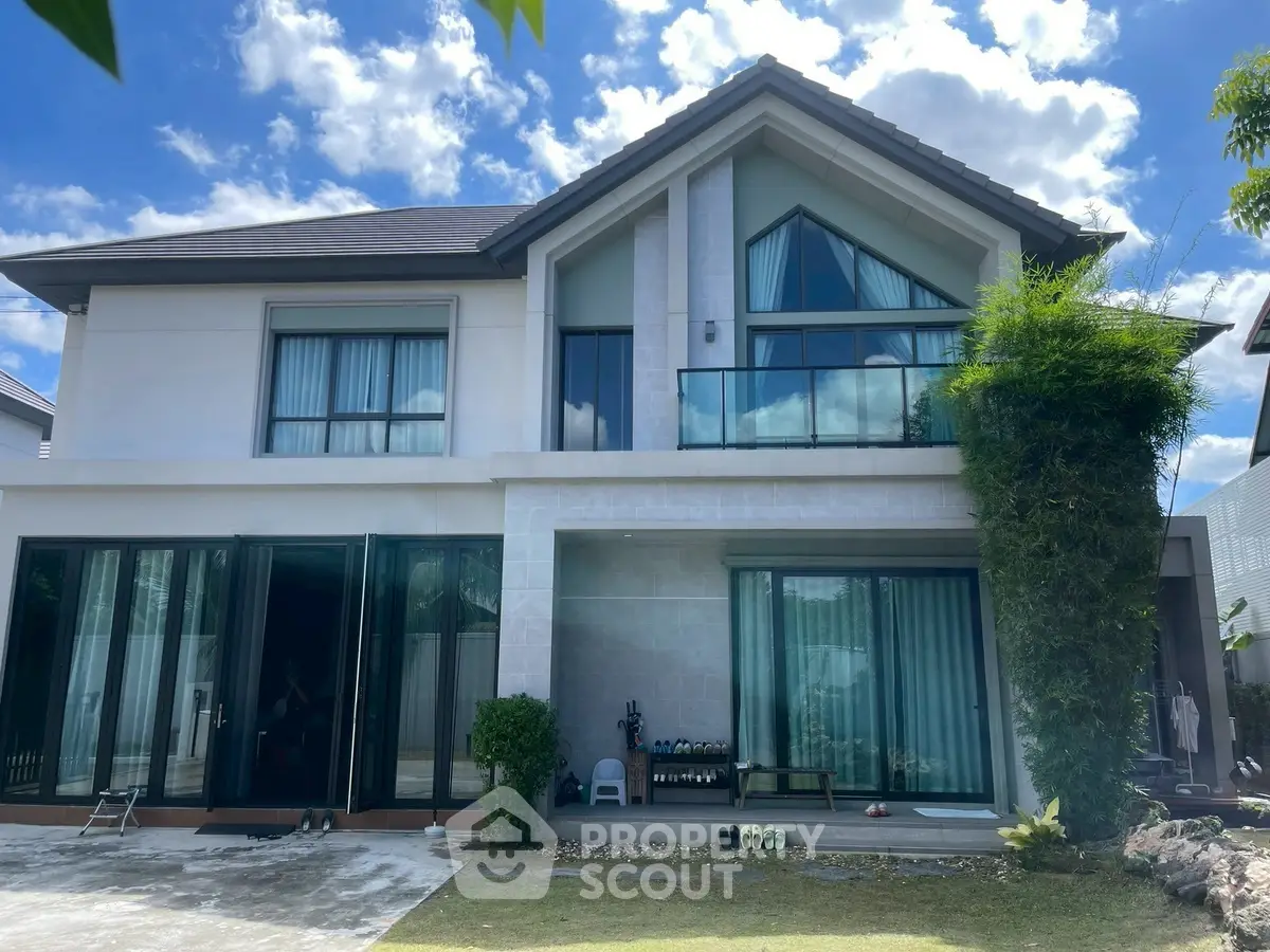 Modern two-story house with large windows and lush greenery under a bright blue sky.
