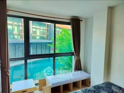 Bright bedroom with large window overlooking pool and modern building, featuring cozy seating area.