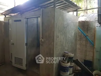Outdoor concrete bathroom with metal roof and door, surrounded by paint buckets and greenery.