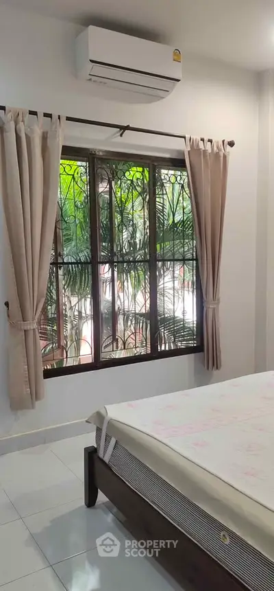 Cozy bedroom with large window and air conditioning, offering a serene garden view.