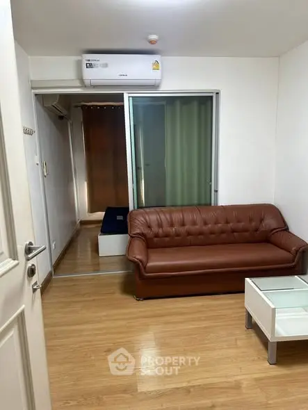 Cozy living room with brown leather sofa and glass coffee table, featuring wooden flooring and air conditioning.
