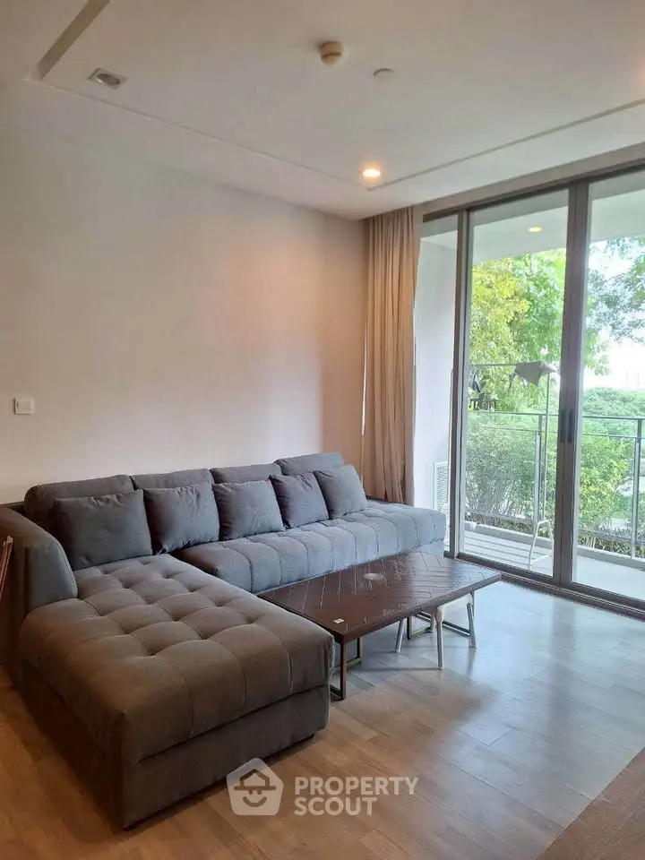 Spacious living room with modern gray sectional sofa and large windows opening to a lush balcony view.