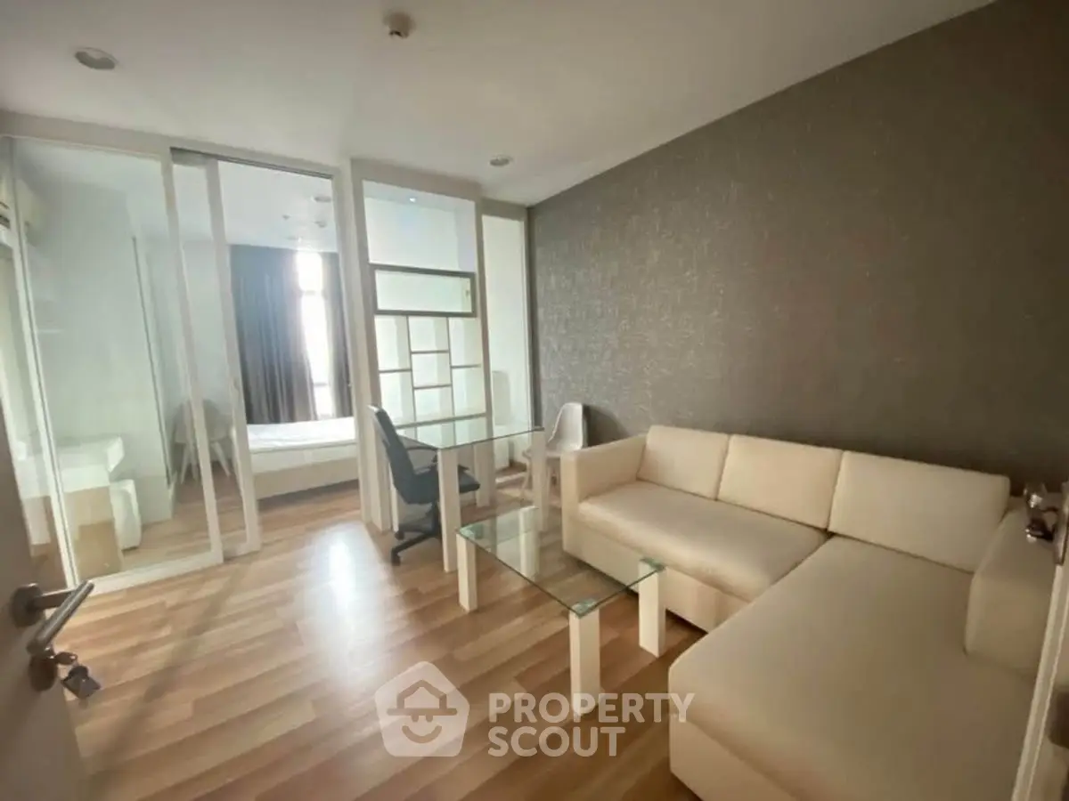 Modern living room with sleek glass table and cozy beige sofa, adjacent to a bright bedroom.