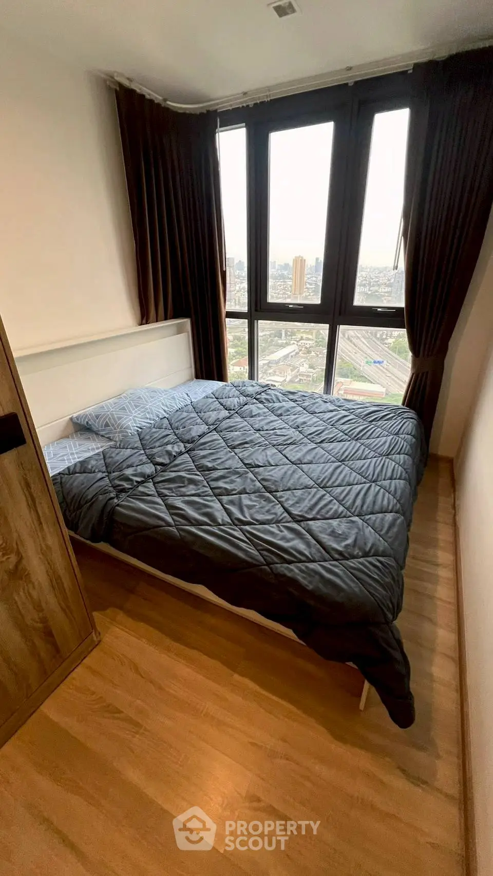 Cozy bedroom with large window offering stunning city views, featuring a comfortable bed and wooden flooring.