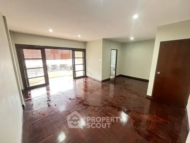 Spacious living room with polished wooden floors and large windows leading to a balcony.