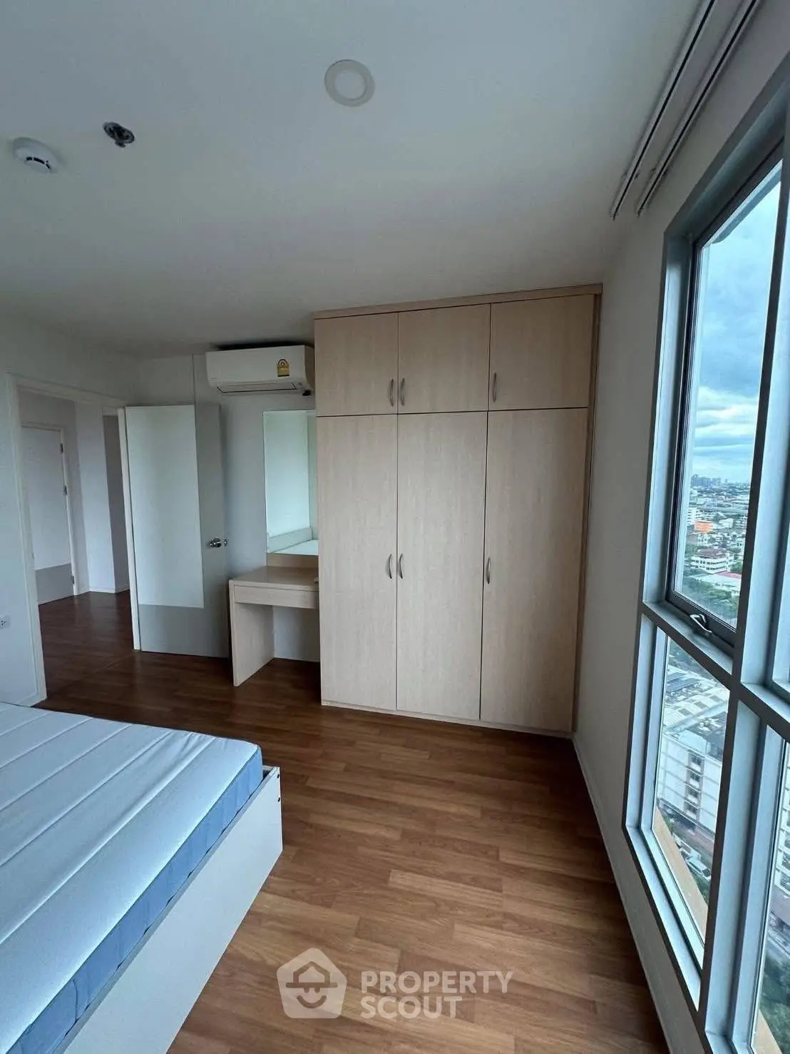 Spacious bedroom with wooden flooring and large window offering city views.