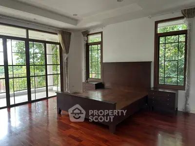 Spacious bedroom with wooden floors and large windows opening to a balcony with lush views.