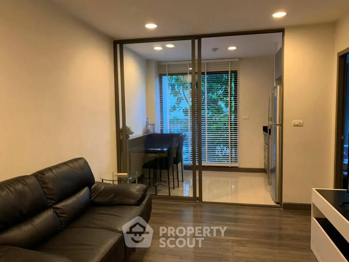 Modern living room with glass partition leading to a bright dining area, featuring sleek furniture and ambient lighting.