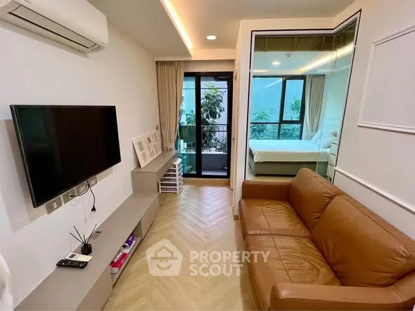 Modern living room with leather sofa and TV, leading to a bedroom with balcony view.