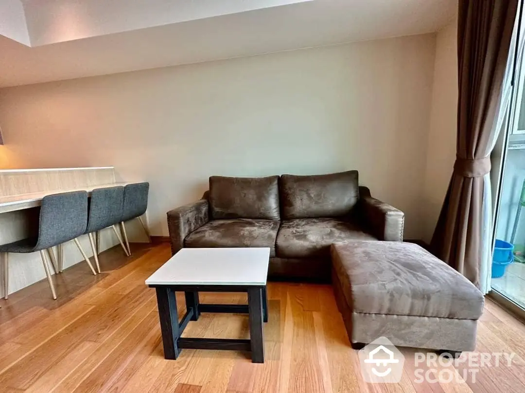 Spacious living room with elegant hardwood flooring, plush brown sectional sofa, and ample natural light from large windows, perfect for relaxation and entertaining guests.