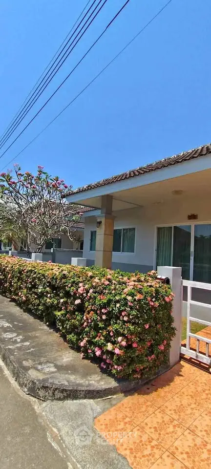 Charming single-story house with blooming garden and tiled pathway under clear blue sky.
