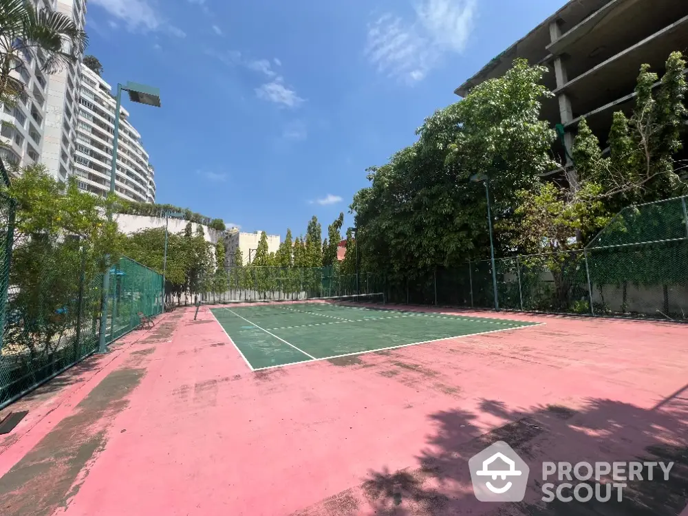 Spacious outdoor tennis court in modern residential complex with lush greenery and clear blue sky.