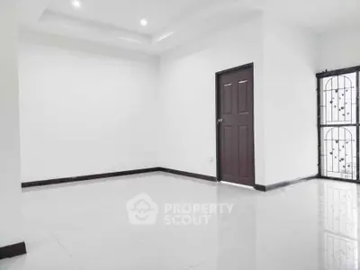 Spacious empty room with white walls and tiled floor, featuring a wooden door and barred windows.