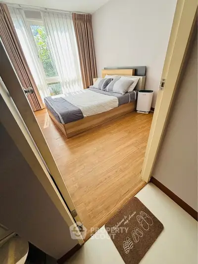 Cozy bedroom with wooden flooring and large window offering natural light, perfect for relaxation.