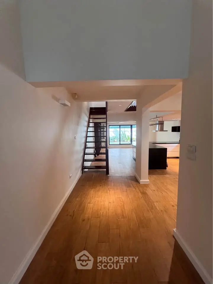 Spacious open-plan living area with wooden flooring and modern staircase, leading to a bright room with large windows.