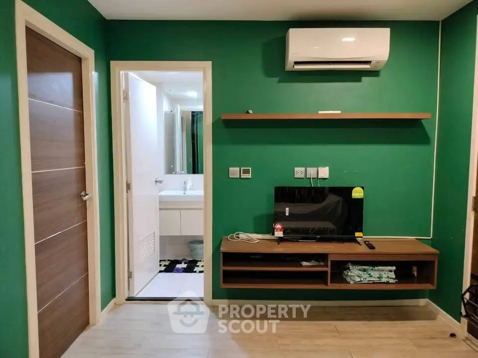 Modern living room with green walls, wall-mounted TV, and air conditioning unit, leading to a bathroom.