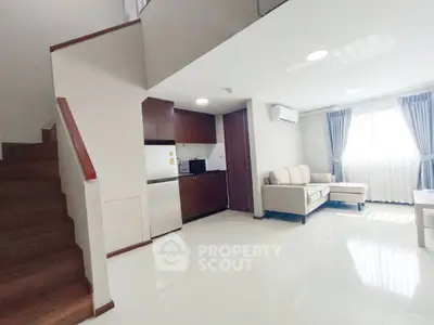 Modern living room with open kitchen and staircase, featuring natural light and cozy seating area.