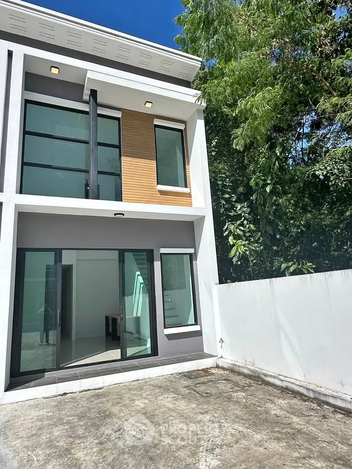 Modern two-story house with large windows and spacious outdoor area, surrounded by lush greenery.