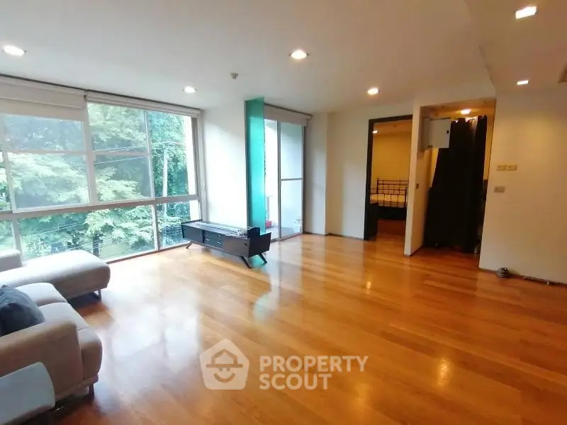 Spacious living room with large windows and wooden flooring, offering a serene view of lush greenery.