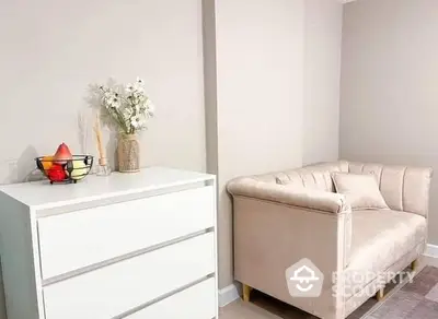 Elegant minimalist living room with a comfortable beige sofa and a chic white dresser, accented by a vase of flowers and a fruit bowl.