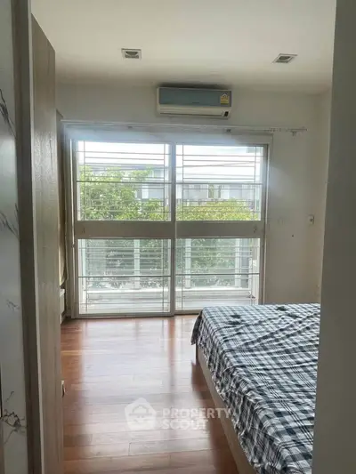 Bright bedroom with large window and air conditioning, featuring a cozy bed and wooden flooring.