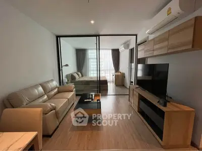 Modern open-plan living room with glass partition leading to bedroom, featuring sleek furniture and natural light.