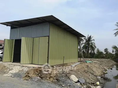 Rural metal shed with open doors on a dirt plot, surrounded by palm trees and a small waterway.