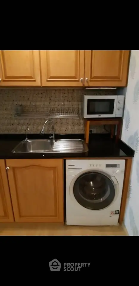 Compact kitchen with wooden cabinets, washing machine, and microwave, ideal for efficient urban living.