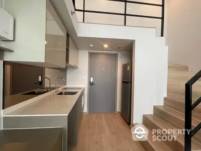 Modern kitchen with sleek countertops and a staircase leading to an upper level, featuring high ceilings and a minimalist design.