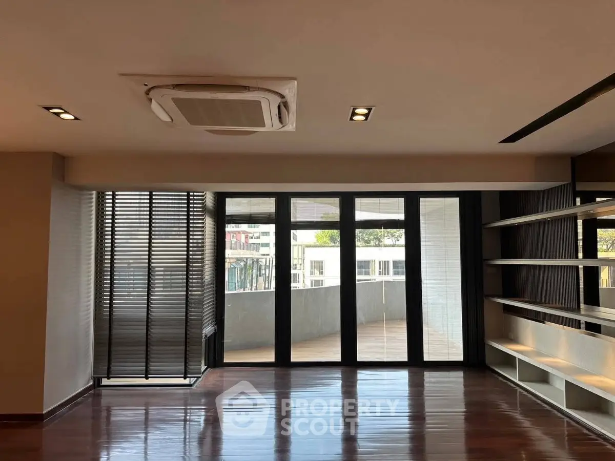 Spacious living room with large windows and balcony access, featuring modern shelving and sleek wooden flooring.