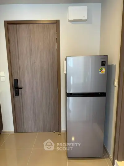 Modern apartment entryway with sleek refrigerator and wooden door, showcasing contemporary design.