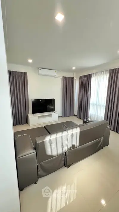 Spacious living room with modern sofa and entertainment center, featuring large windows and elegant curtains.