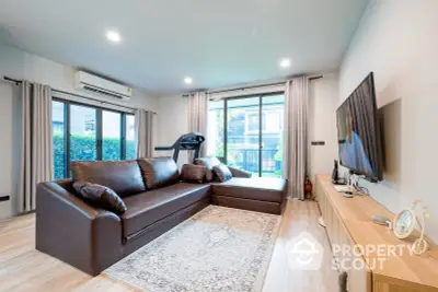 Spacious living room with modern decor, large sectional sofa, and treadmill, opening to a sunny balcony with greenery.