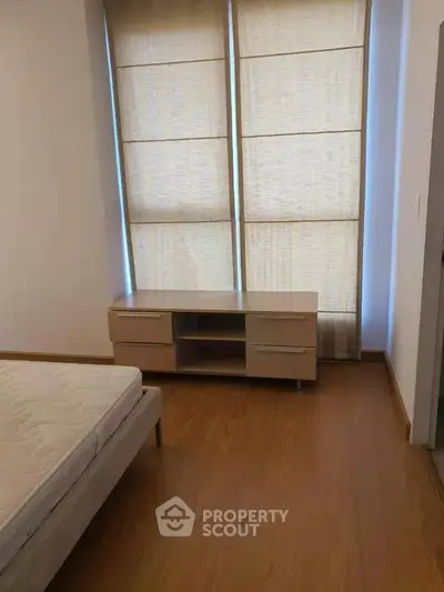 Spacious bedroom with wooden flooring and modern furniture, featuring large windows with blinds.