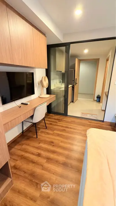Modern bedroom with wooden flooring and study area, featuring a sleek kitchen view through glass doors.