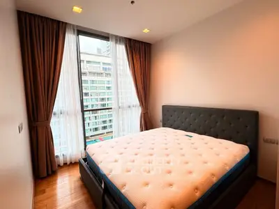 Modern bedroom with large window and city view, featuring a stylish bed and elegant curtains.