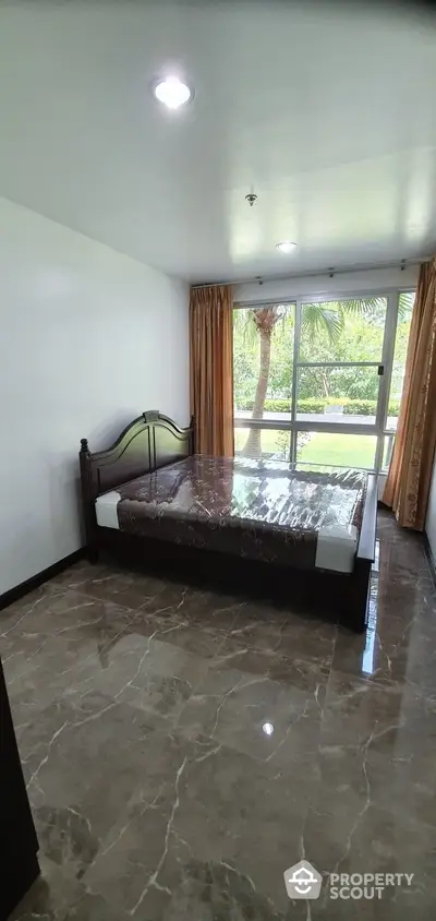 Spacious bedroom with polished marble flooring, large windows offering lush garden views, and a classic wooden bed frame.