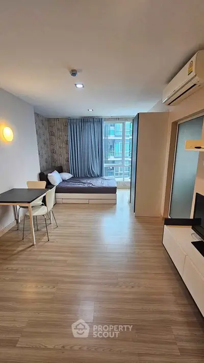 Modern studio apartment with wooden flooring, cozy bed, and dining area, featuring large windows for natural light.