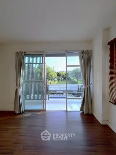 Spacious living room with large windows and balcony overlooking lush greenery, perfect for relaxation.