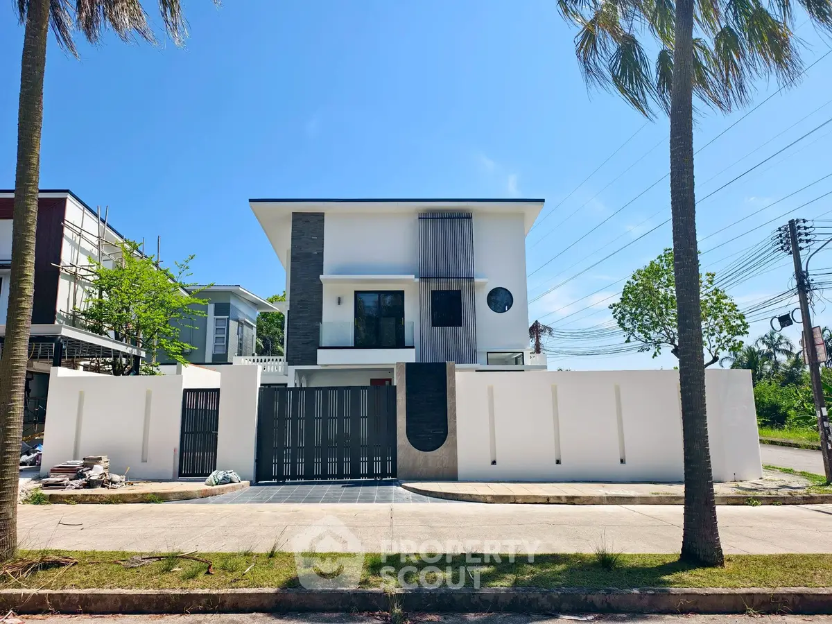 Modern two-story house with sleek design and gated entrance, surrounded by palm trees and clear blue sky.