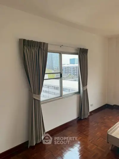 Bright bedroom with large window and elegant curtains, showcasing city view and polished wooden floor.